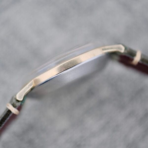 A close-up side view of the Wittnauer Date Manual Wind 2732-1-C189, highlighting its metal case and leather strap at the edge and lugs, set against a textured gray background. The watch face is not shown.