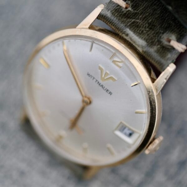 A close-up of the Wittnauer Date Manual Wind 2732-1-C189 wristwatch, featuring a gold case, white dial with gold hour markers, date window at 3 o’clock, and a dark leather strap. The watch face displays the time as 5:12.