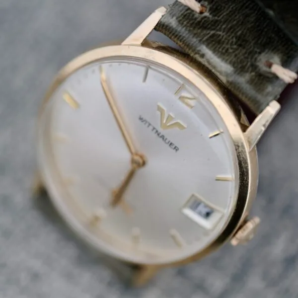 A close-up of the Wittnauer Date Manual Wind 2732-1-C189 wristwatch, featuring a gold case, white dial with gold hour markers, date window at 3 o’clock, and a dark leather strap. The watch face displays the time as 5:12.