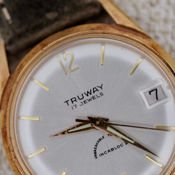 Close-up of a vintage Truway 17 Jewels Date Incabloc wristwatch with a gold case, white face, gold hour markers, black hands, “17 jewels” inscription, and a date window displaying the number 7.