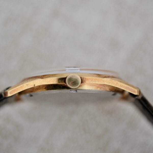 A close-up side view of the Truway 17 Jewels Date Incabloc wristwatch showcases its gold-toned case, round crown, and black strap on a light textured surface. The watch face is not visible.