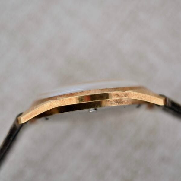 Close-up side view of the Truway 17 Jewels Date Incabloc wristwatch case in gold with a black strap, highlighting its flat, angular profile and brushed finish against a neutral, textured background.