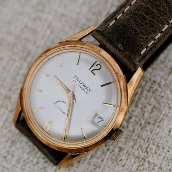 A close-up photo of a Truway 17 Jewels Date Incabloc wristwatch with a gold-tone case, white dial, date window set at 17, and a worn brown leather strap, displayed on a textured light surface.