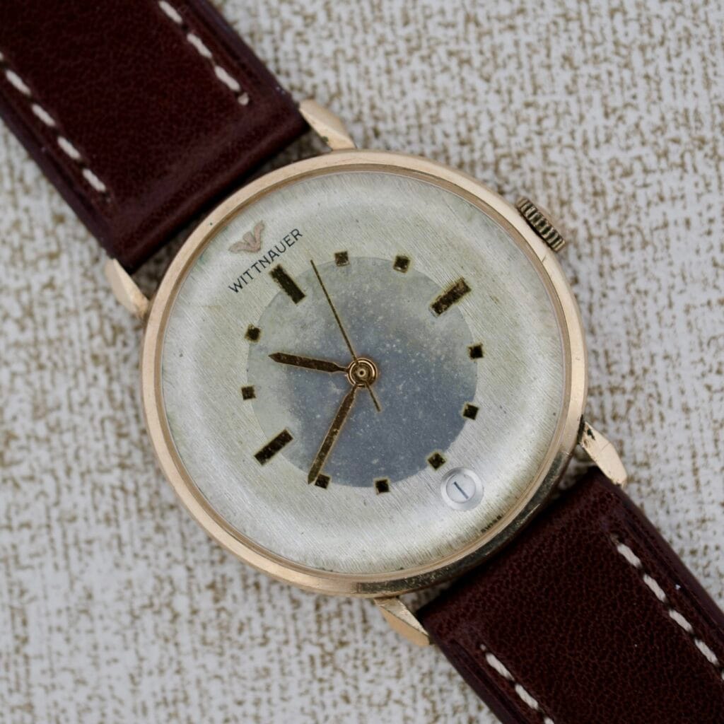 Wittnauer 627-1-215 Manual Wind Date wristwatch with a brown leather strap and date display, displayed on a textured surface.