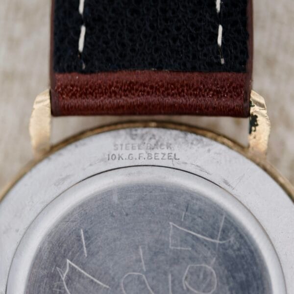 Close-up of the Wittnauer 627-1-215 Manual Wind Date case back engraved with STEEL BACK 10K G.F. BEZEL, showing a black strap featuring brown leather accents.