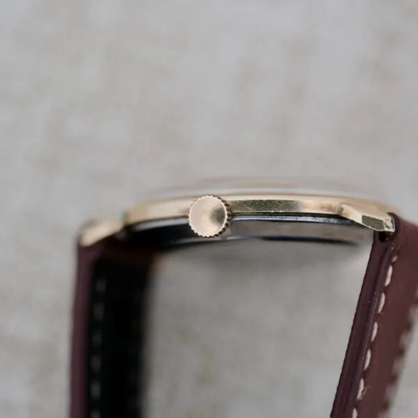 Close-up side view of the Wittnauer 627-1-215 Manual Wind Date wristwatch, featuring a gold-colored crown, thin case, and brown leather strap with white stitching, set against a light textured background.