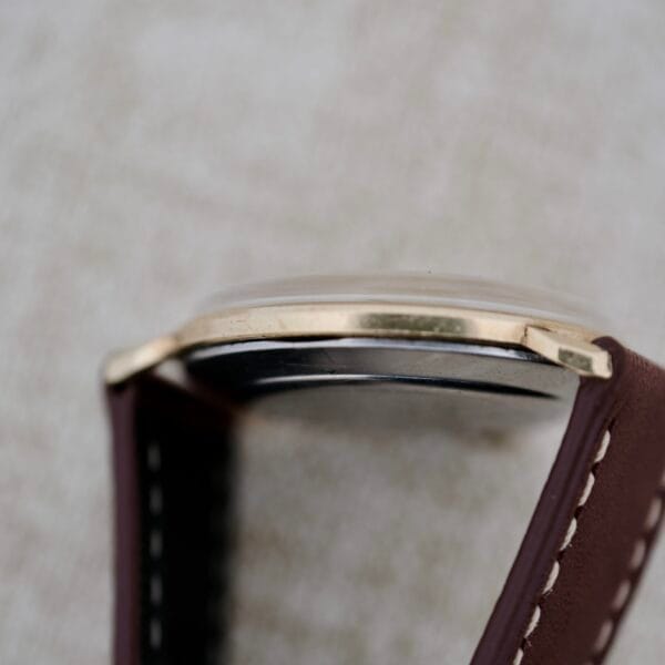 Close-up side view of the Wittnauer 627-1-215 Manual Wind Date wristwatch featuring a brown leather strap with white stitching, gold-tone bezel, and slightly domed glass set against a softly blurred background.