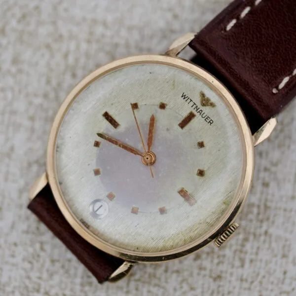 A Wittnauer 627-1-215 Manual Wind Date wristwatch with a gold case, worn off-white dial, gold markers and hands, date window, and brown leather strap with white stitching is shown on a textured surface.