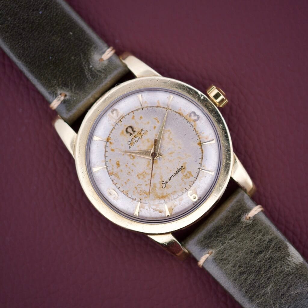 An Omega Seamaster Automatic 2577-17 Tropic Dial watch with a worn dial and dark leather strap rests on a maroon surface.