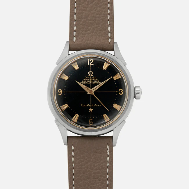 A wristwatch with a round black dial, gold hour markers and hands, silver casing, and a brown leather strap. The dial features the Omega logo, Automatic Chronometer, Constellation text, and references the classic Omega Constellation 2782.