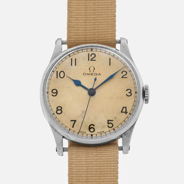 A vintage Omega wristwatch with a beige face, black numerals, blue hands, and a tan fabric strap. The watch shows slight wear and patina, indicating age. The Omega logo is visible below the 12 o’clock position.