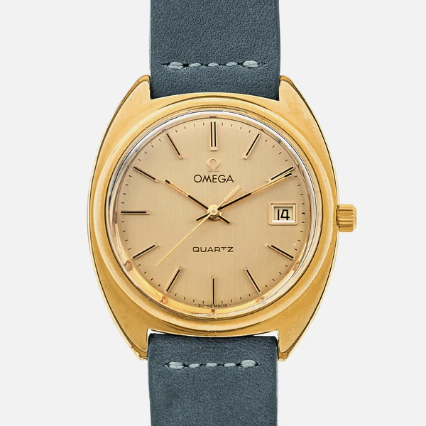 A gold Omega Quartz wristwatch, ref. Omega 196.0121, featuring a round beige dial, gold hands and hour markers, a date window at 3 o’clock, and a black leather strap. The watch crown sits on the right side.