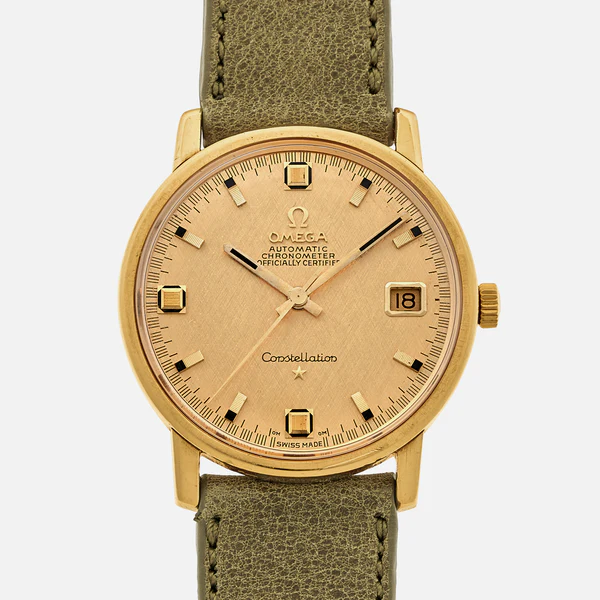 A gold Omega Constellation automatic chronometer wristwatch with a beige dial, date window at 3 oclock, gold hour markers, and a green leather strap is shown against a plain background.