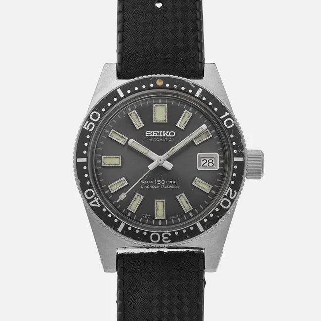 A Seiko automatic wristwatch featuring a black dial, seiko branding, rectangular hour markers, a date window at 3 o’clock, rotating bezel, and black textured strap. The face displays Water 150m Proof and Diashock 17 Jewels.