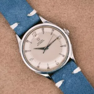 An Omega Automatic Bumper 2635 Cal. 351 wristwatch with a blue strap rests on a tan surface, displaying the time as 6:40.