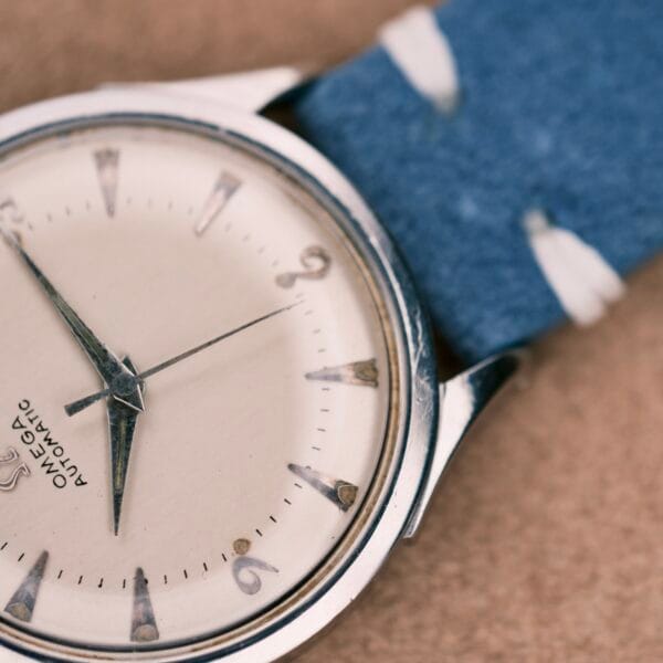 Close-up of an Omega Automatic Bumper 2635 Cal. 351 wristwatch with a blue strap and silver hour markers, displayed on a beige surface.
