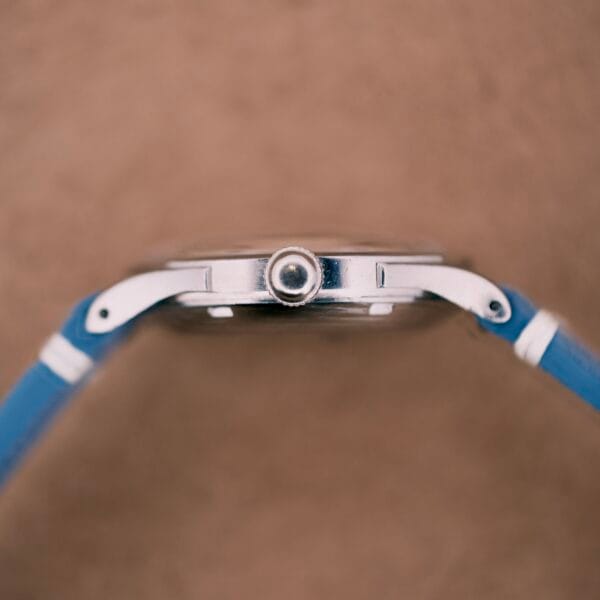 Side view of an Omega Automatic Bumper 2635 Cal. 351 wristwatch with a blue strap and silver case, set against a beige background.
