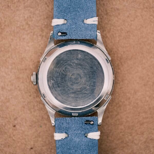 The back of an Omega Automatic Bumper 2635 Cal. 351 silver wristwatch with a blue leather strap rests on a tan surface.