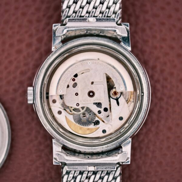 Close-up of the Movado Automatic 431 Stainless Steel watch movement, showcasing its visible gears and engraved details against a brown textured background.