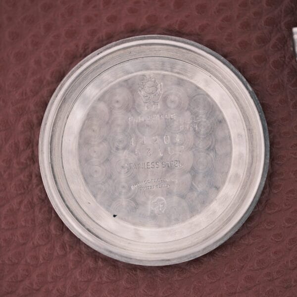 A round Movado Automatic 431 Stainless Steel watch back with engraved text, resting on a textured maroon surface.