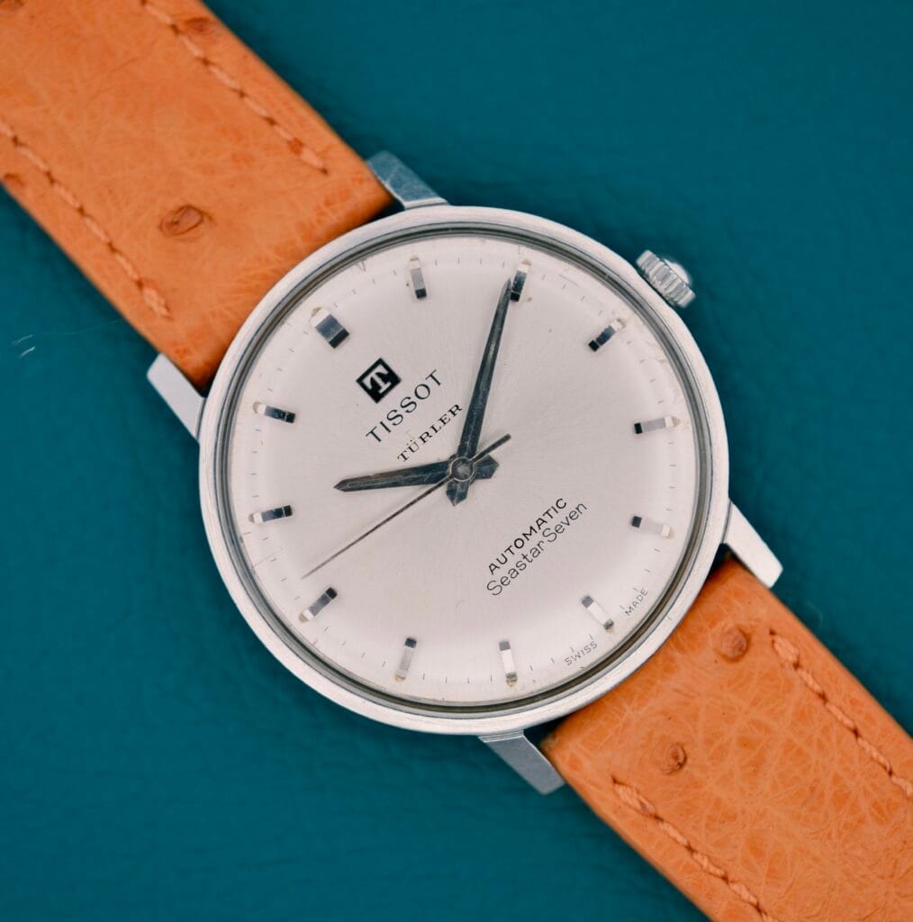 A Tissot Automatic Seastar Seven Turler wristwatch with a tan leather strap is shown on a teal background, displaying the time 11:09.