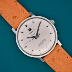 A Tissot Automatic Seastar Seven Turler wristwatch with a tan leather strap is shown on a teal background, displaying the time 11:09.