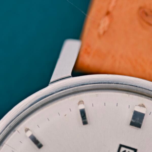 Close-up of a Tissot Automatic Seastar Seven Turler silver watch face with a beige leather strap, set against a teal background.