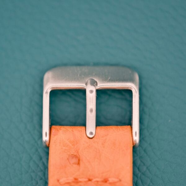 Close-up of a silver buckle on a tan leather strap, the Tissot Automatic Seastar Seven Turler detail stands out against a textured teal background.