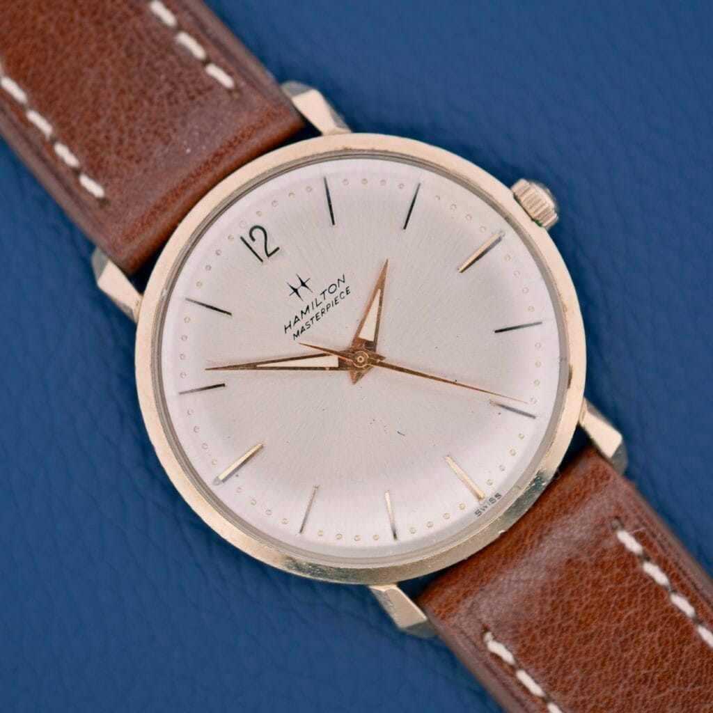 A Hamilton Stormking VII Masterpiece wristwatch with a brown leather strap rests on a blue textured surface.