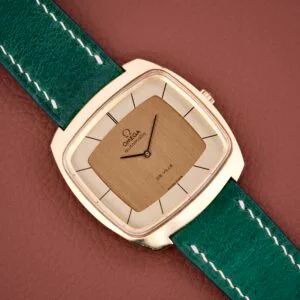 The Omega Deville 151.0051 TV Case watch features a square gold design with a green leather strap, showcased on a brown textured surface.