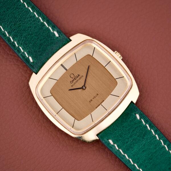 The Omega Deville 151.0051 TV Case watch features a square gold design with a green leather strap, showcased on a brown textured surface.