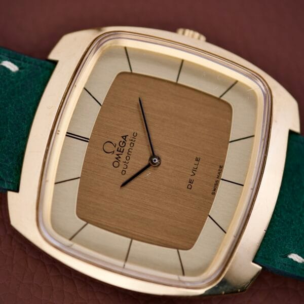 The Omega Deville 151.0051 TV Case watch features a square face, green strap, and black hands, displayed on a brown textured surface.