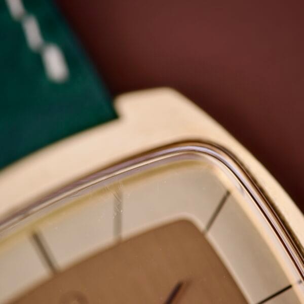 Close-up of the Omega Deville 151.0051 TV Case, showcasing its unique watch face and glass, gold casing, and part of a green strap against a brown background.