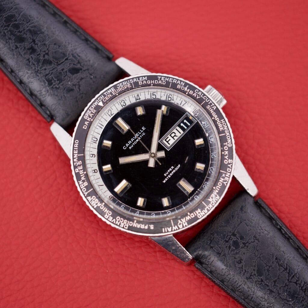 A Caravelle Worldtimer Ref. 753 Day/Date with a black dial and black leather strap rests on a red surface.