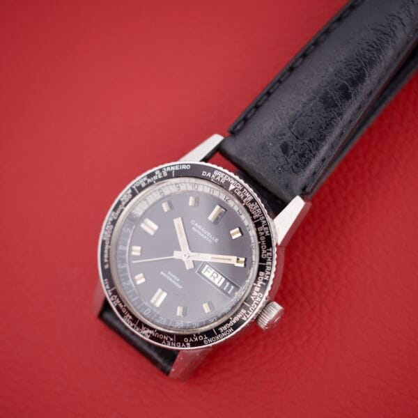 A Caravelle Worldtimer Ref. 753 Day/Date wristwatch with a black leather strap is displayed on a textured red surface.