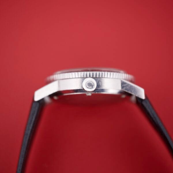 Side view of the Caravelle Worldtimer Ref. 753 Day/Date wristwatch with a black strap set against a red background, highlighting the crown and day/date display.