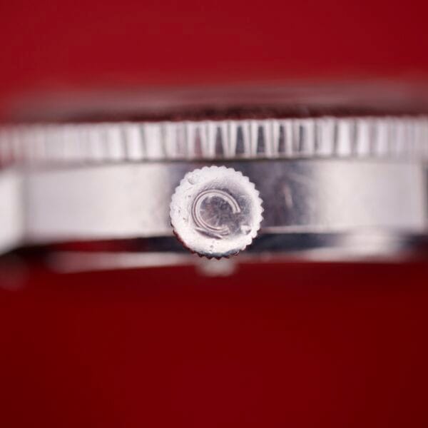 Close-up of the Caravelle Worldtimer Ref. 753 Day/Date watch, highlighting its silver crown, bezel, and day/date display against a solid red background.