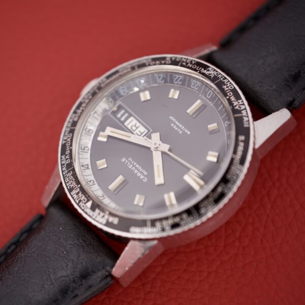 Close-up of a vintage Caravelle Worldtimer Ref. 753 Day/Date wristwatch with a black leather strap, displayed on a red textured background.