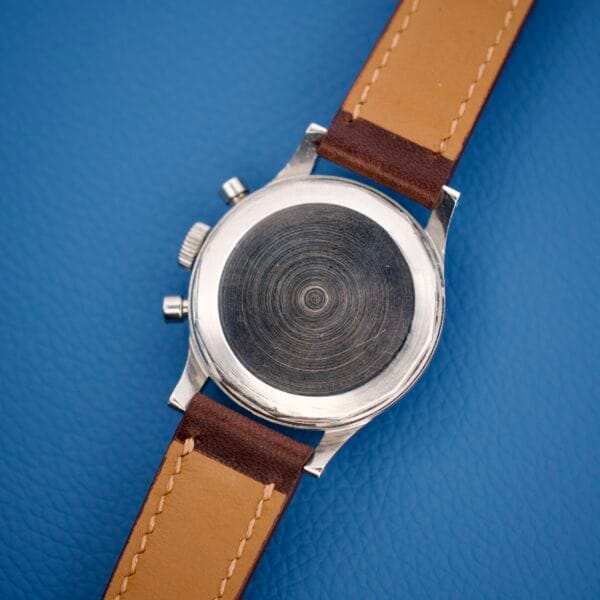 The Wittnauer Chronograph Venus 188, featuring a blank metal case back and brown leather strap, is displayed on a blue textured surface, highlighting its classic movement style.