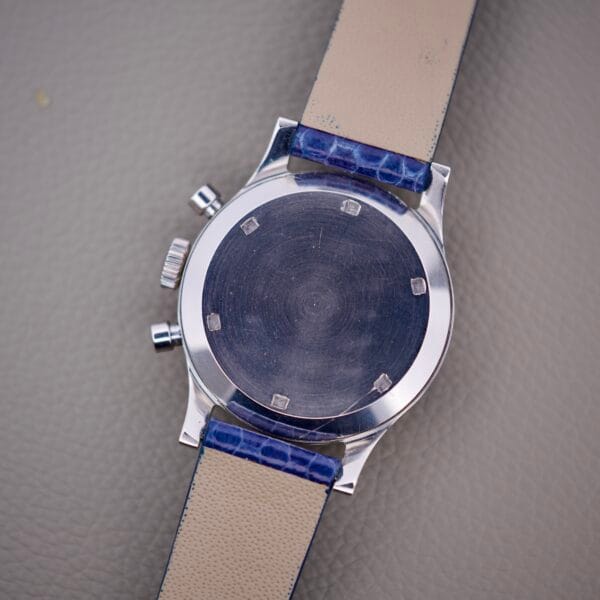 A Wittnauer 3256 Chronograph Venus 188 wristwatch with a blank metal case back and blue leather strap, displayed face down on a gray textured surface.