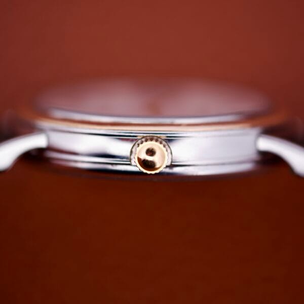 Close-up side view of a Girard Perregaux Automatic 4797 silver wristwatch with a gold crown, set against a blurred brown background.