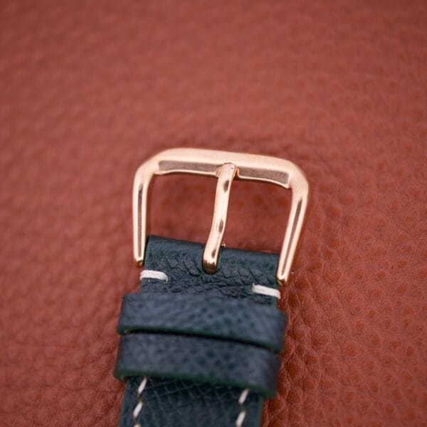 A close-up of a Girard Perregaux Automatic 4797 gold buckle on a black leather strap, set against a textured brown background.