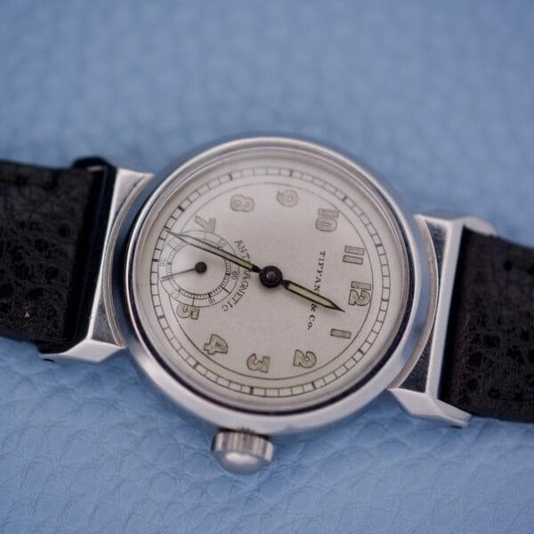 The Tiffany & Co. By Movado 11729 silver vintage wristwatch with a black leather strap is elegantly displayed on a light blue textured surface.