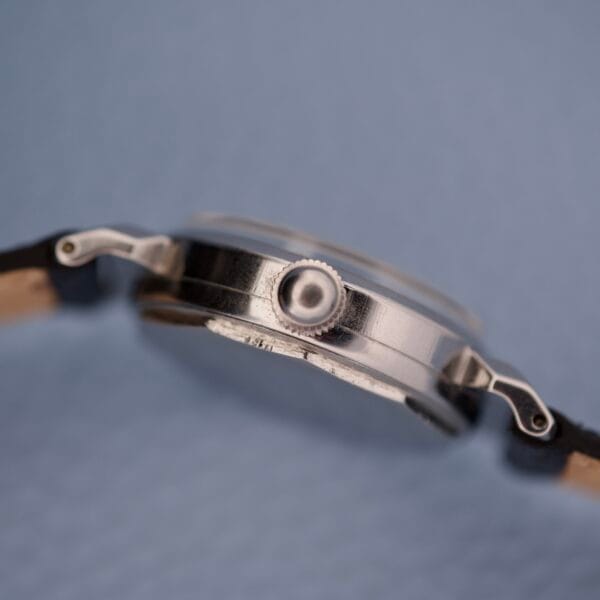 Side view of the Tiffany & Co. By Movado 11729, a sleek silver wristwatch with a black strap, focused on the crown against a light blue background.