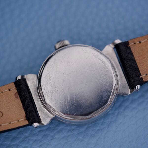 A close-up of the scratched back of a Tiffany & Co. By Movado 11729 silver wristwatch with a black and tan leather strap, resting on a blue surface.