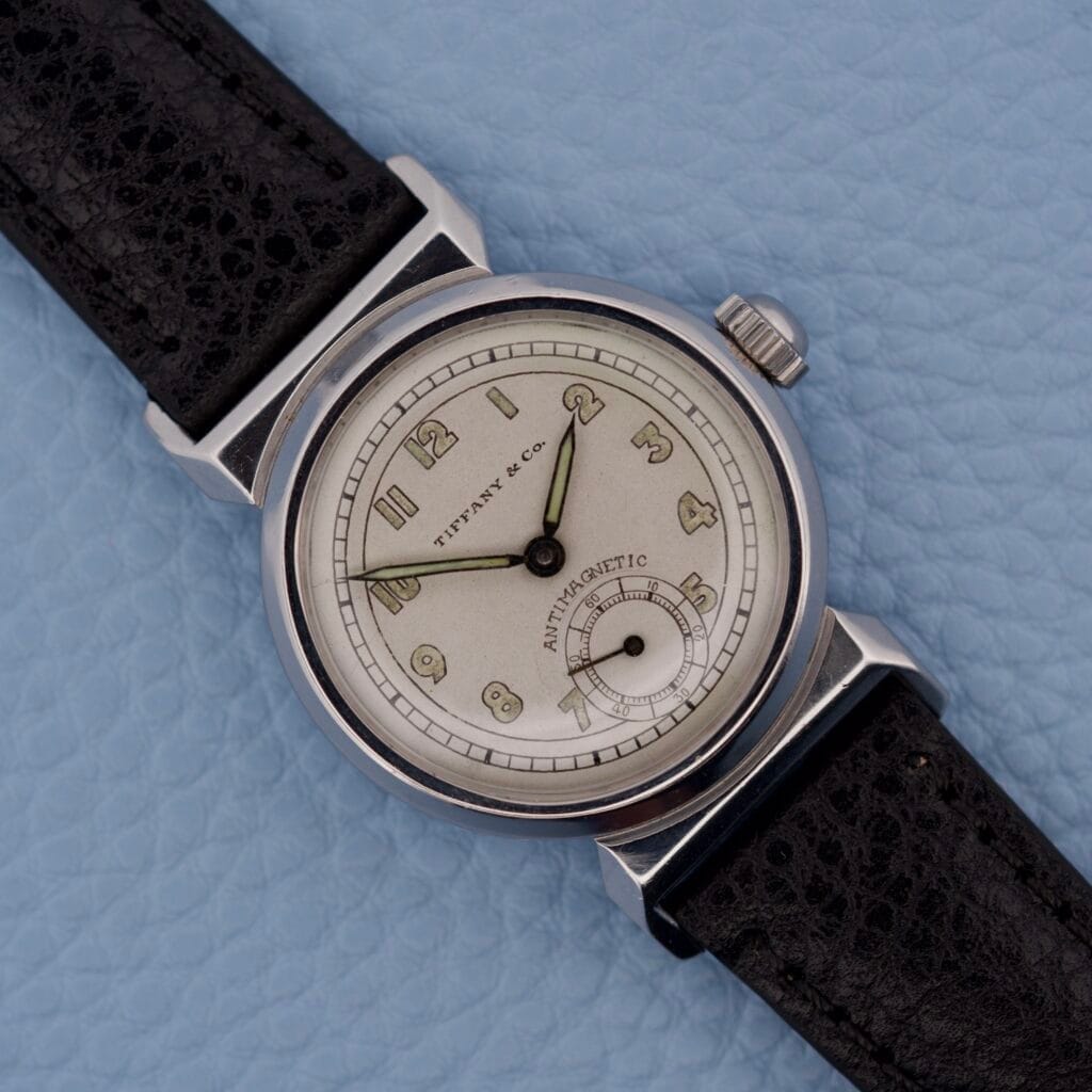 A Tiffany & Co. By Movado 11729 wristwatch with a black leather strap rests on a light blue textured surface.