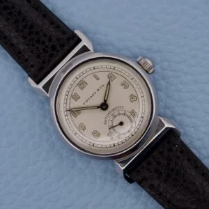 A Tiffany & Co. By Movado 11729 wristwatch with a black leather strap rests on a light blue textured surface.