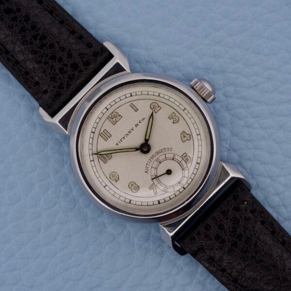 A Tiffany & Co. By Movado 11729 wristwatch with a black leather strap rests on a light blue textured surface.