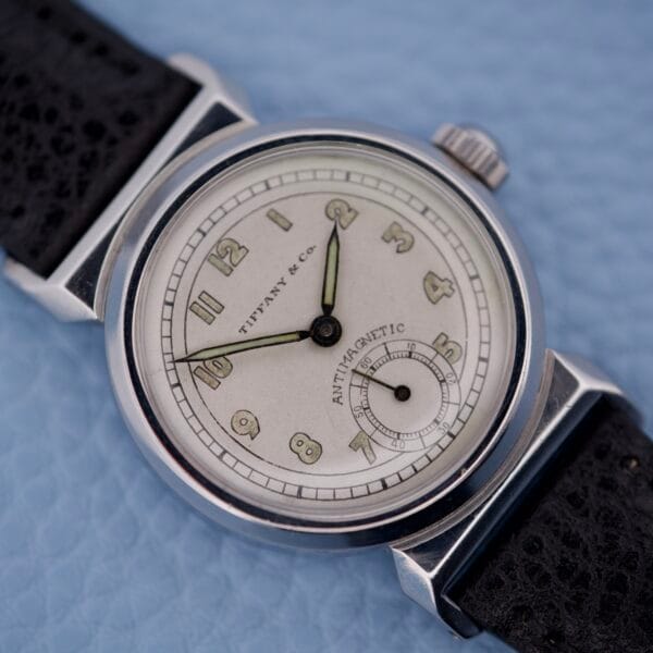 Close-up of a Tiffany & Co. By Movado 11729 vintage wristwatch with a black strap, resting on a light blue textured surface.
