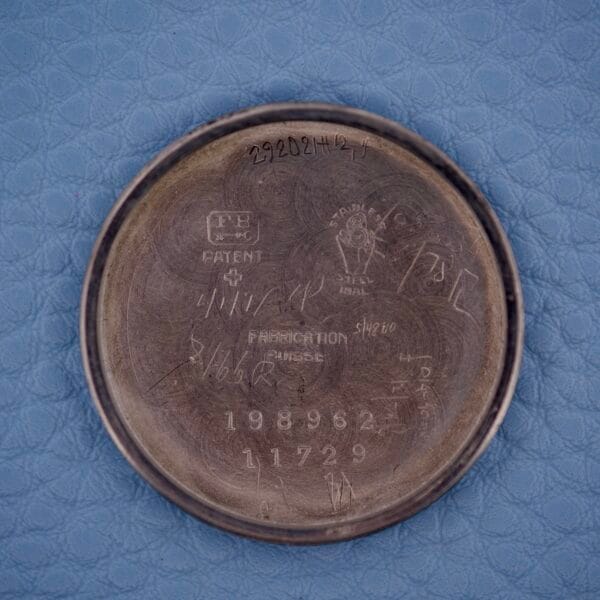 The inside case back of a Tiffany & Co. By Movado 11729 watch displays various engraved numbers and markings, shown on a blue textured surface.